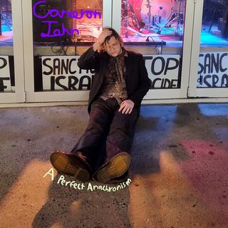 Cameron John sitting on the sidewalk outside of the venue. The name "Cameron John" is handwritten out on the window, and the title "A Perfect Anachronism" at his feet. Perspective has been corrected for viewers in the Northern hemisphere